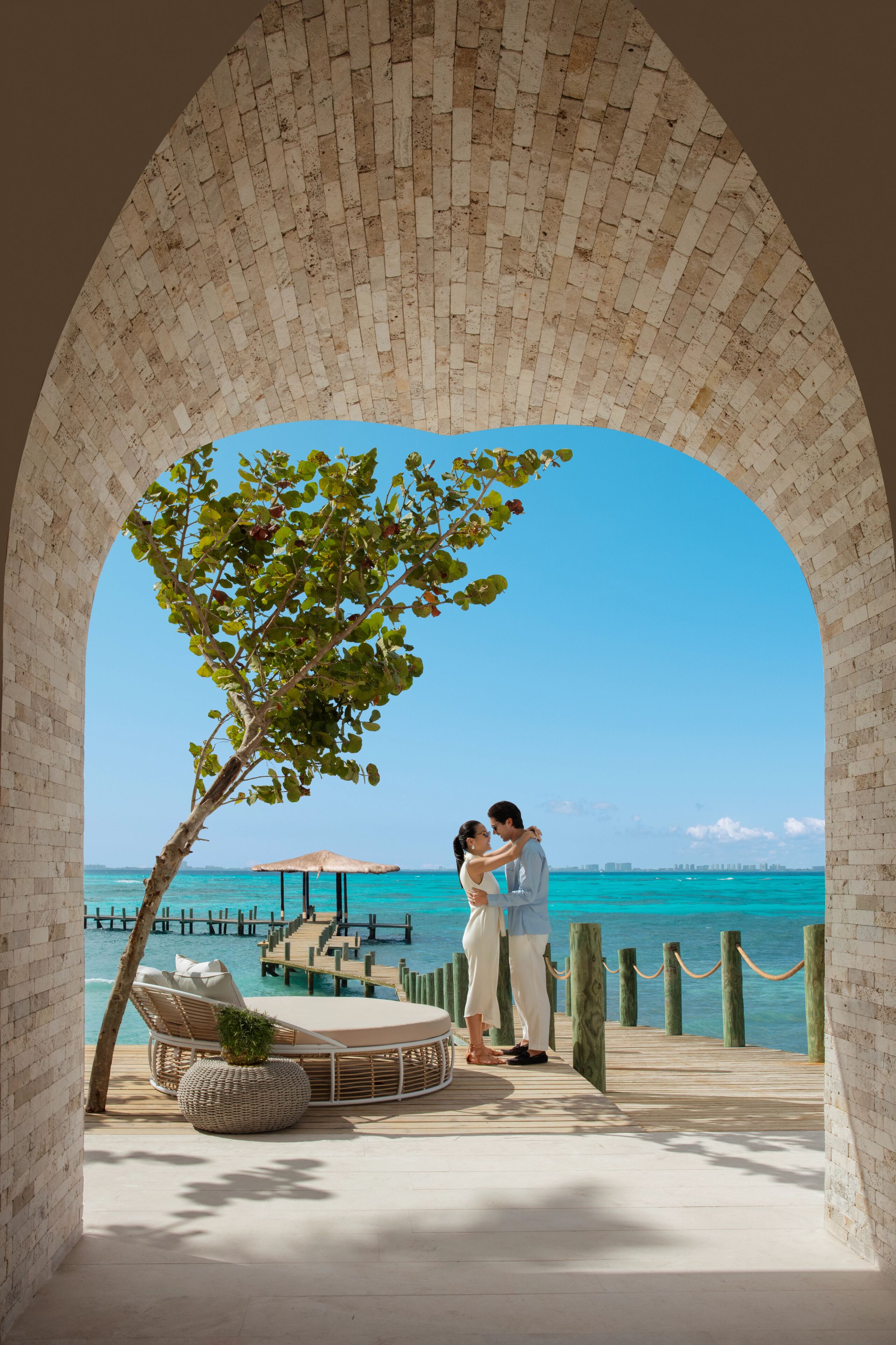 A couple embracing under a brick archway overlooking a beach with a wooden pier and lounge chairs.