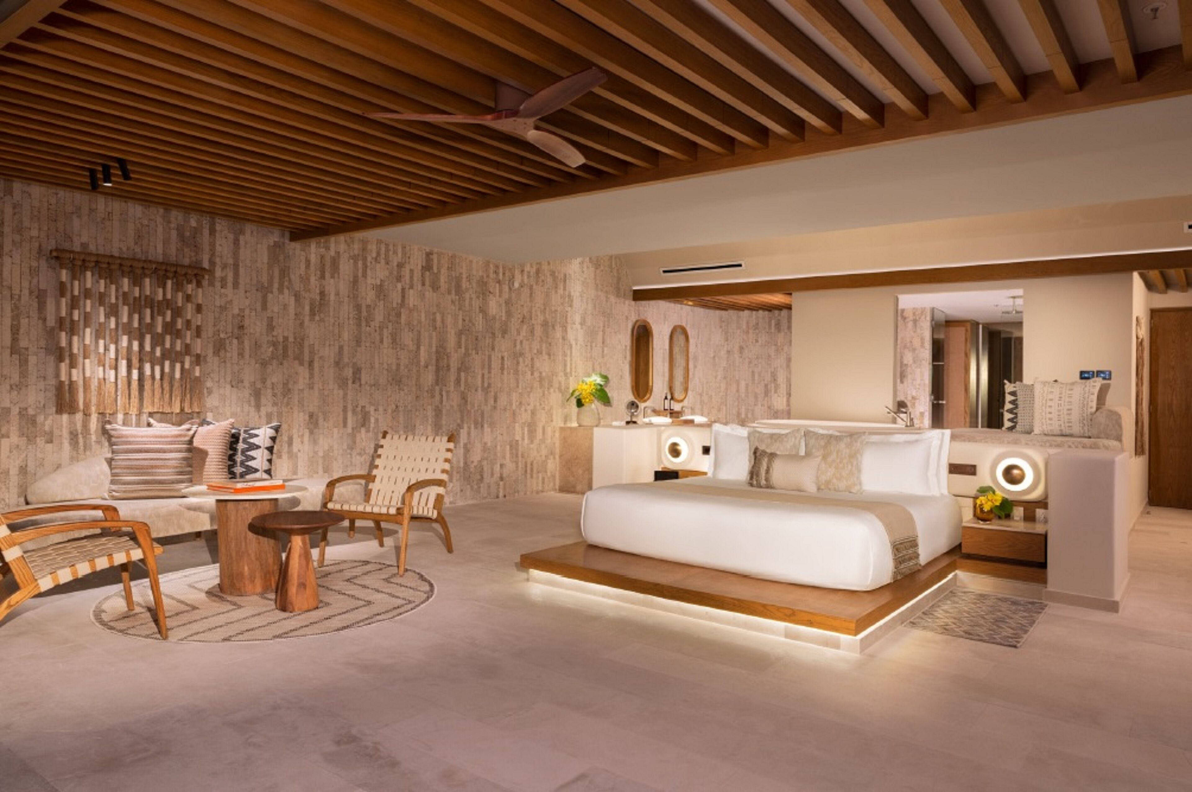 Een moderne slaapkamer met een houten plafond, een groot bed, een nachtkastje, een spiegel en een zithoek met een bank en fauteuils.