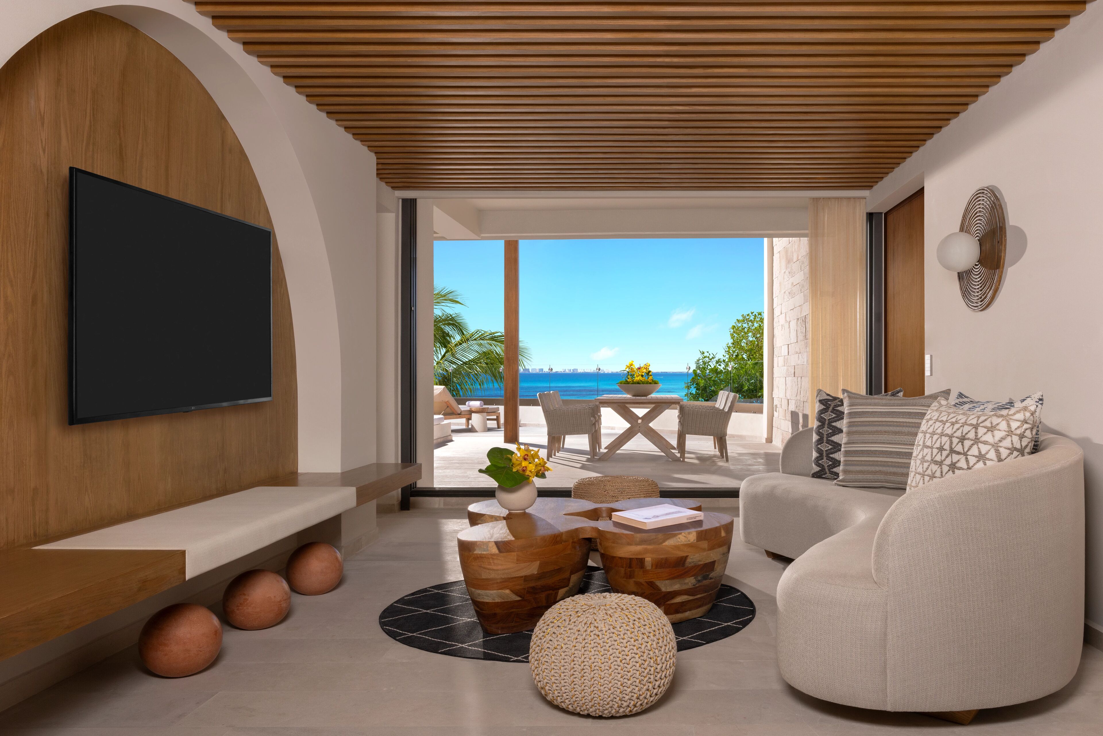 A modern living room with a large TV, wooden furniture, and a view of the ocean.