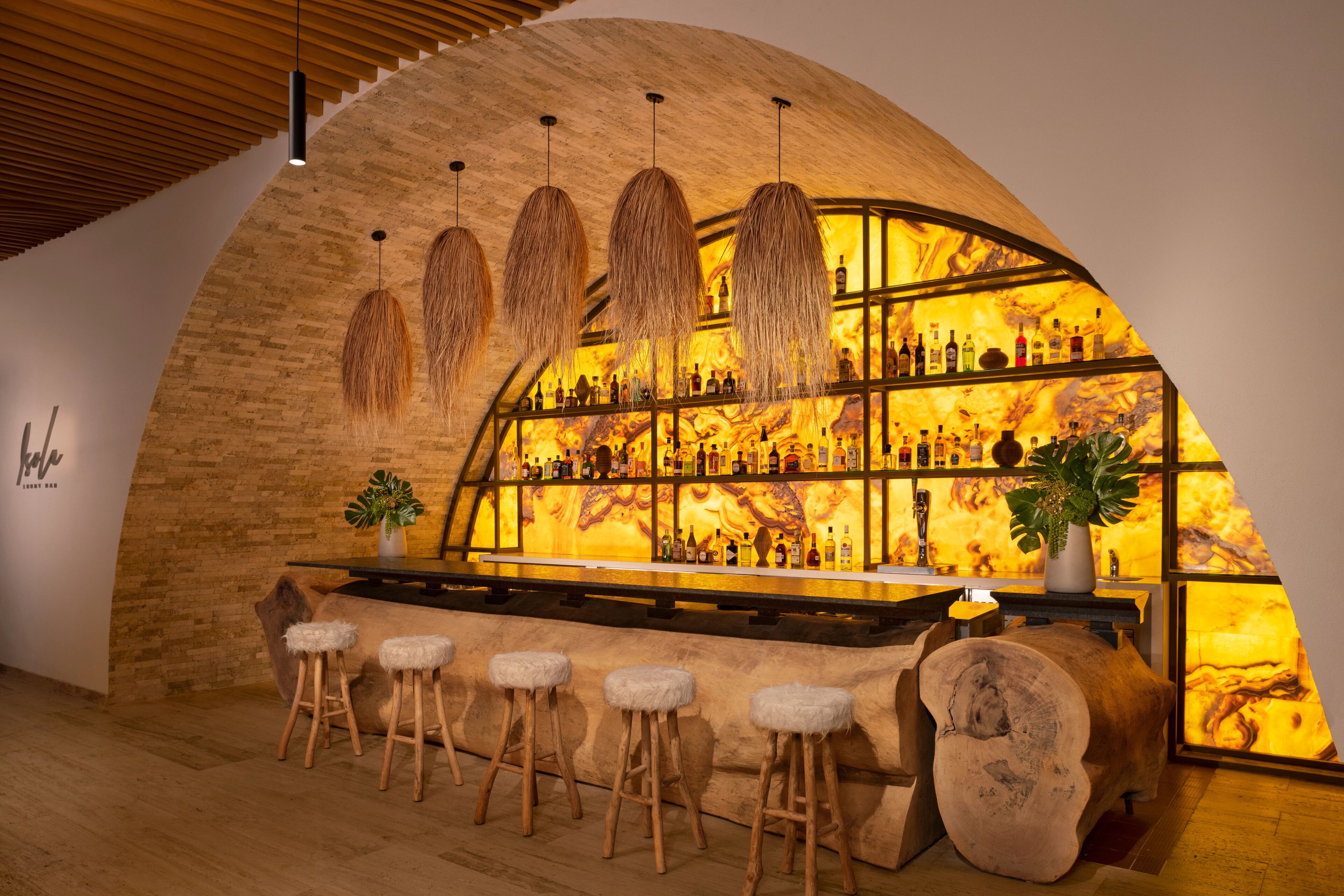 Un bar con techo de madera curvada, luces colgantes y una estantería llena de botellas. Hay taburetes y un tronco grande como adorno.