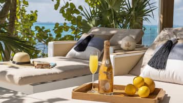 A sunlit lounge area with a view of the sea, featuring a bottle of champagne, glasses, and lemons on a wooden tray.