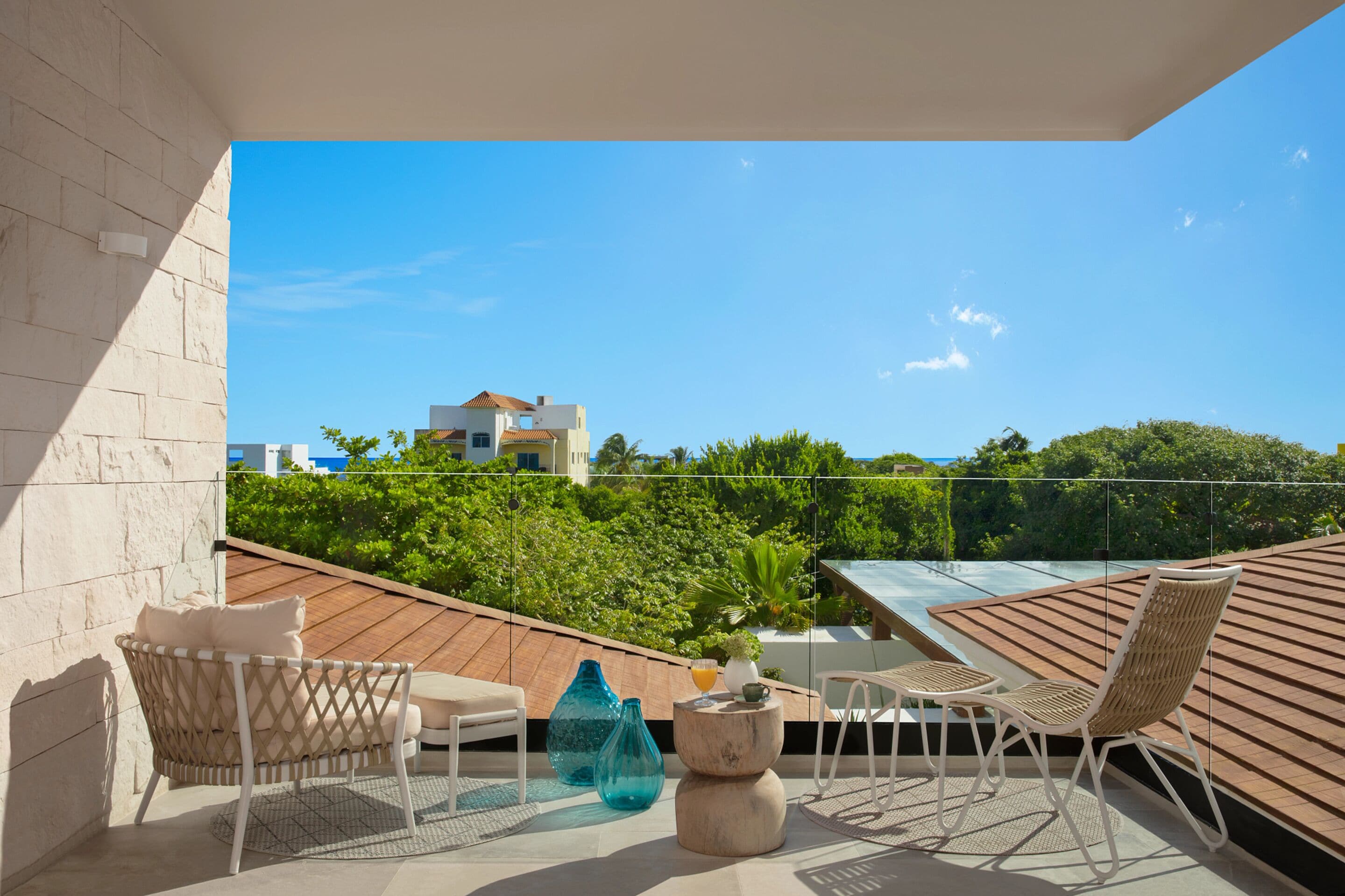 Un balcón con sillones de mimbre, una mesita y barandilla de vidrio con vistas a un jardín exuberante y un edificio en la distancia.