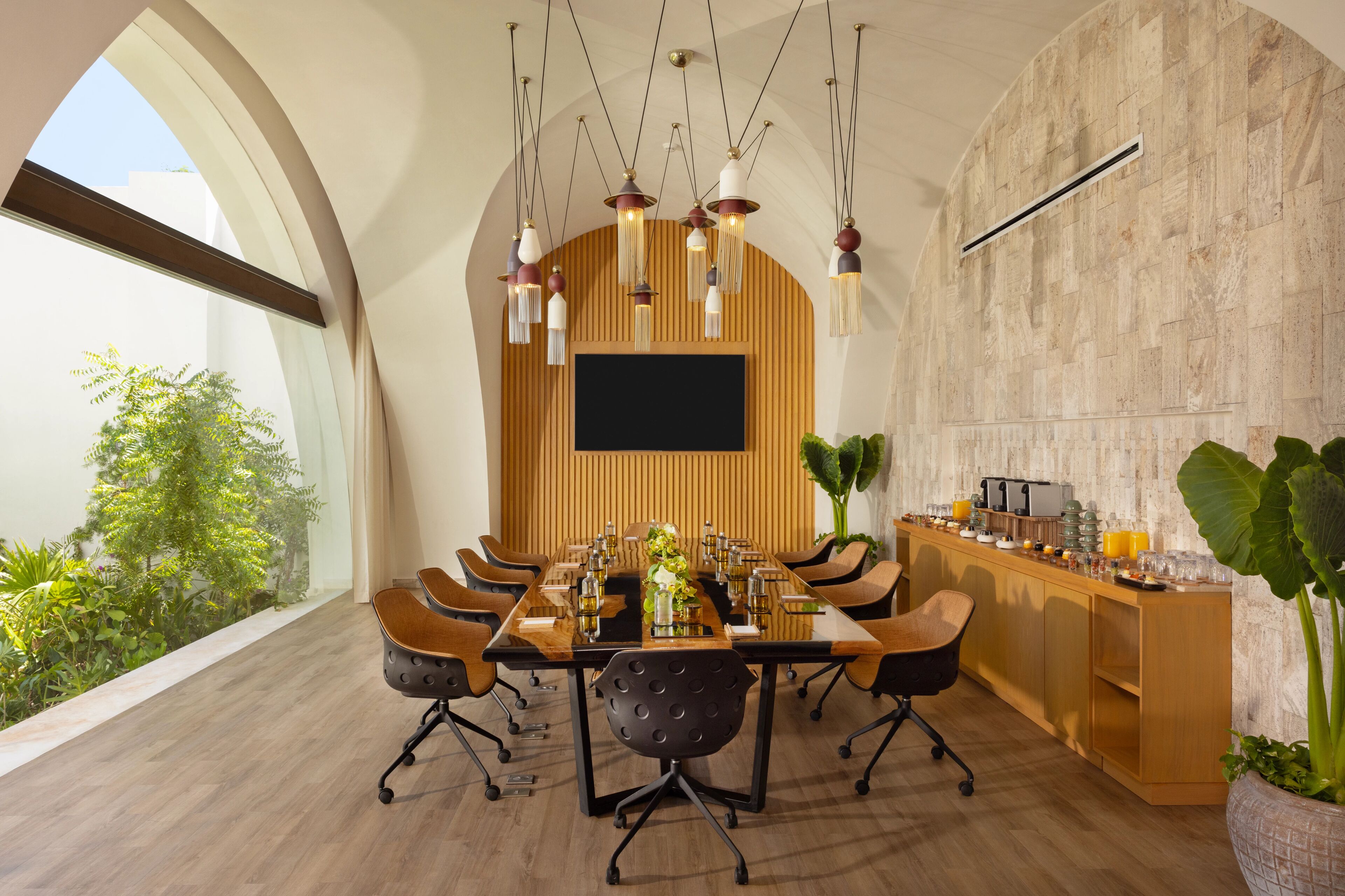 A modern conference room with a long table, multiple chairs, hanging pendant lights, a flat-screen TV, and a wooden accent wall.
