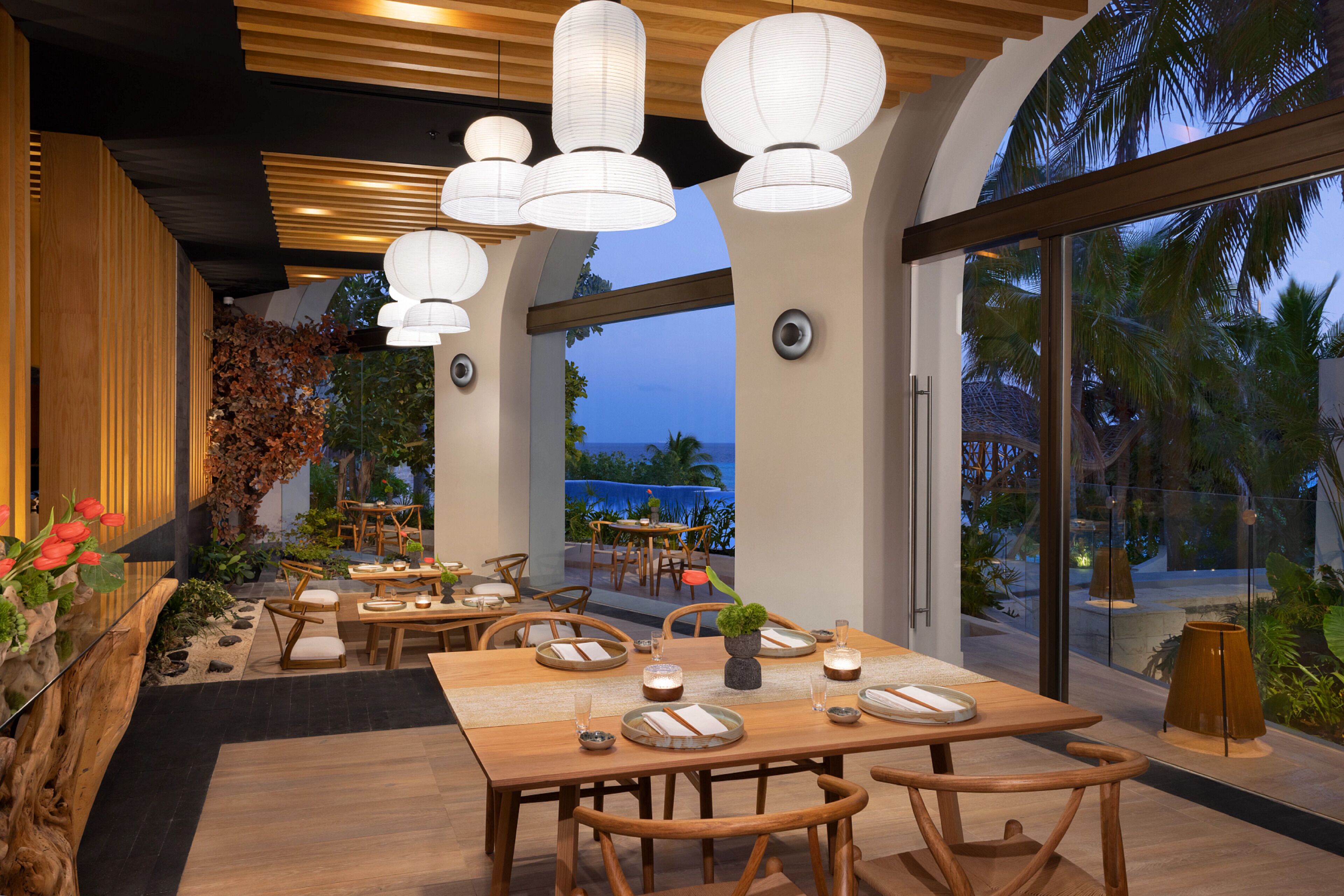 A restaurant with wooden tables and chairs, white paper lanterns, and a view of the ocean at dusk.