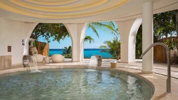 A pool area with clear water, a view of the ocean, and a tropical setting with palm trees.