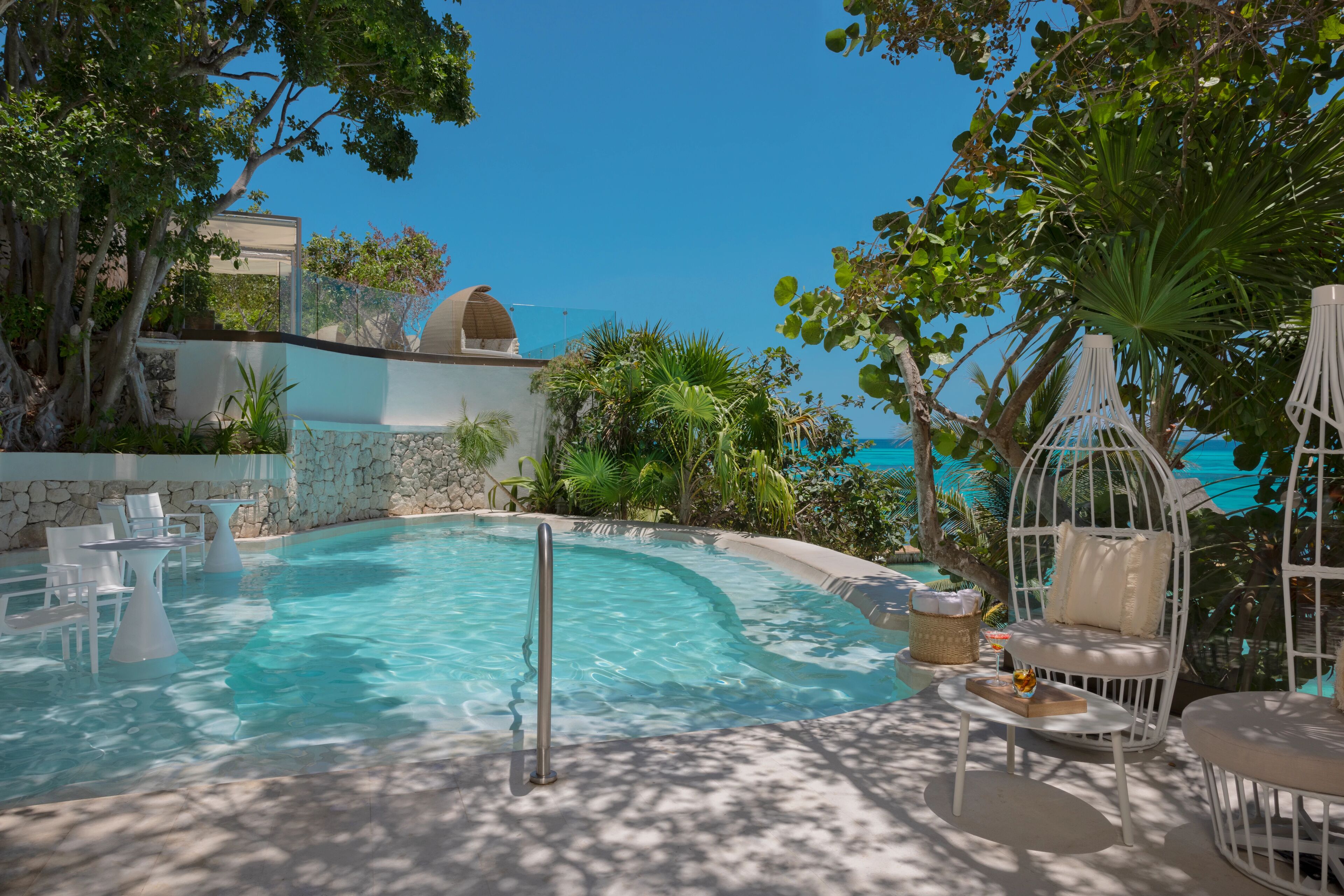A pool area with a curved edge, surrounded by white lounge chairs and tables, and a view of the ocean.