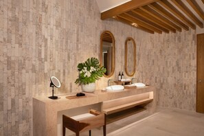 A modern bathroom with a stone wall, two sinks, a mirror, a bench, and wooden beams on the ceiling.