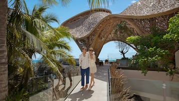 Un couple se promène sur une passerelle aux murs de verre, d’où l’on peut admirer la mer et une structure architecturale unique, notamment son toit tressé.