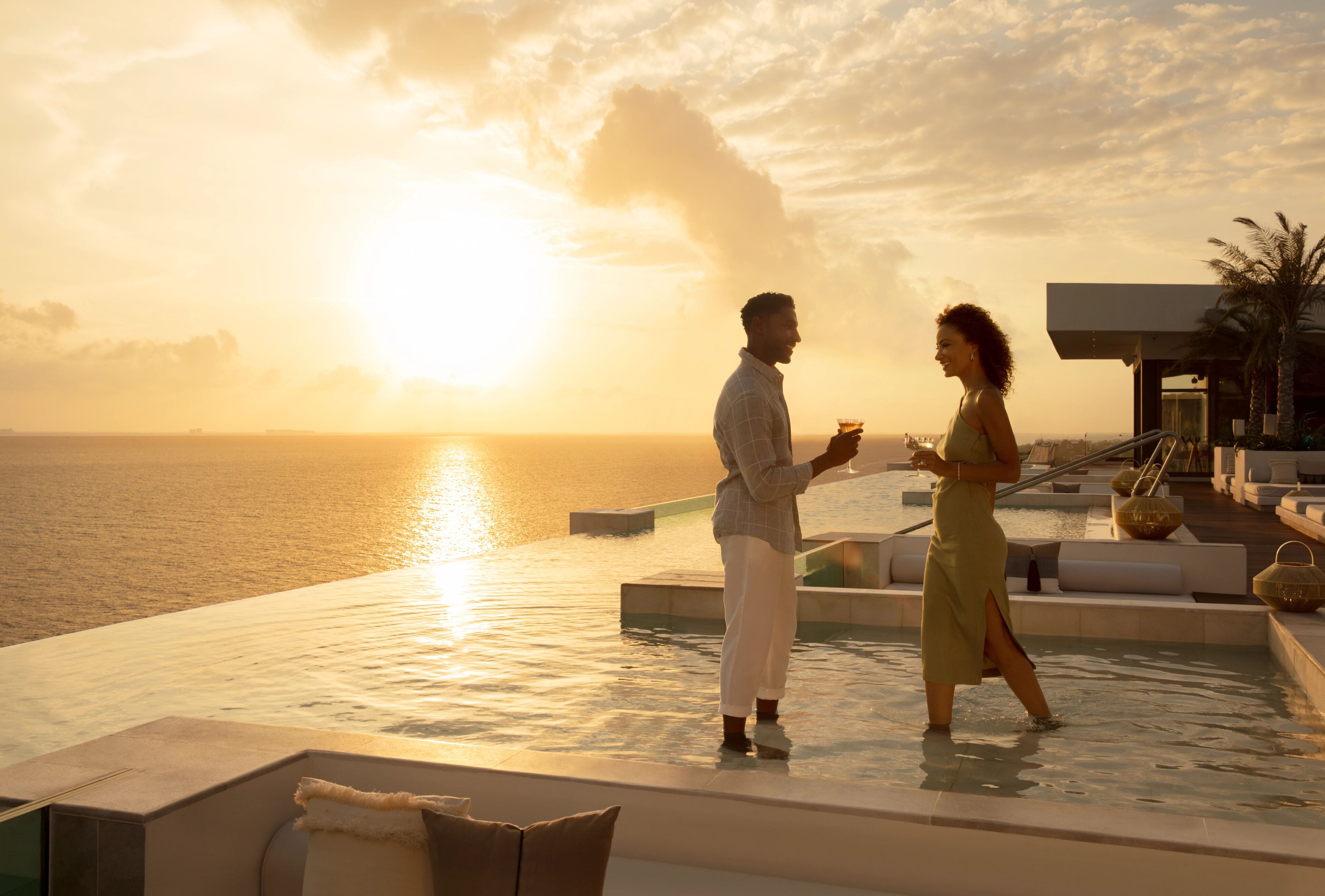 Una pareja parada junto a una piscina infinita al atardecer, con vista al océano.