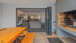 Decoração moderna com mesa de madeira e bancos, sofá cinza e área de cozinha visível através de uma porta aberta.