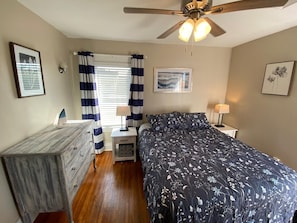 Une chambre à coucher comprenant un lit, une commode, des tables de chevet, des lampes, un ventilateur de plafond et des œuvres d’art accrochées aux murs.