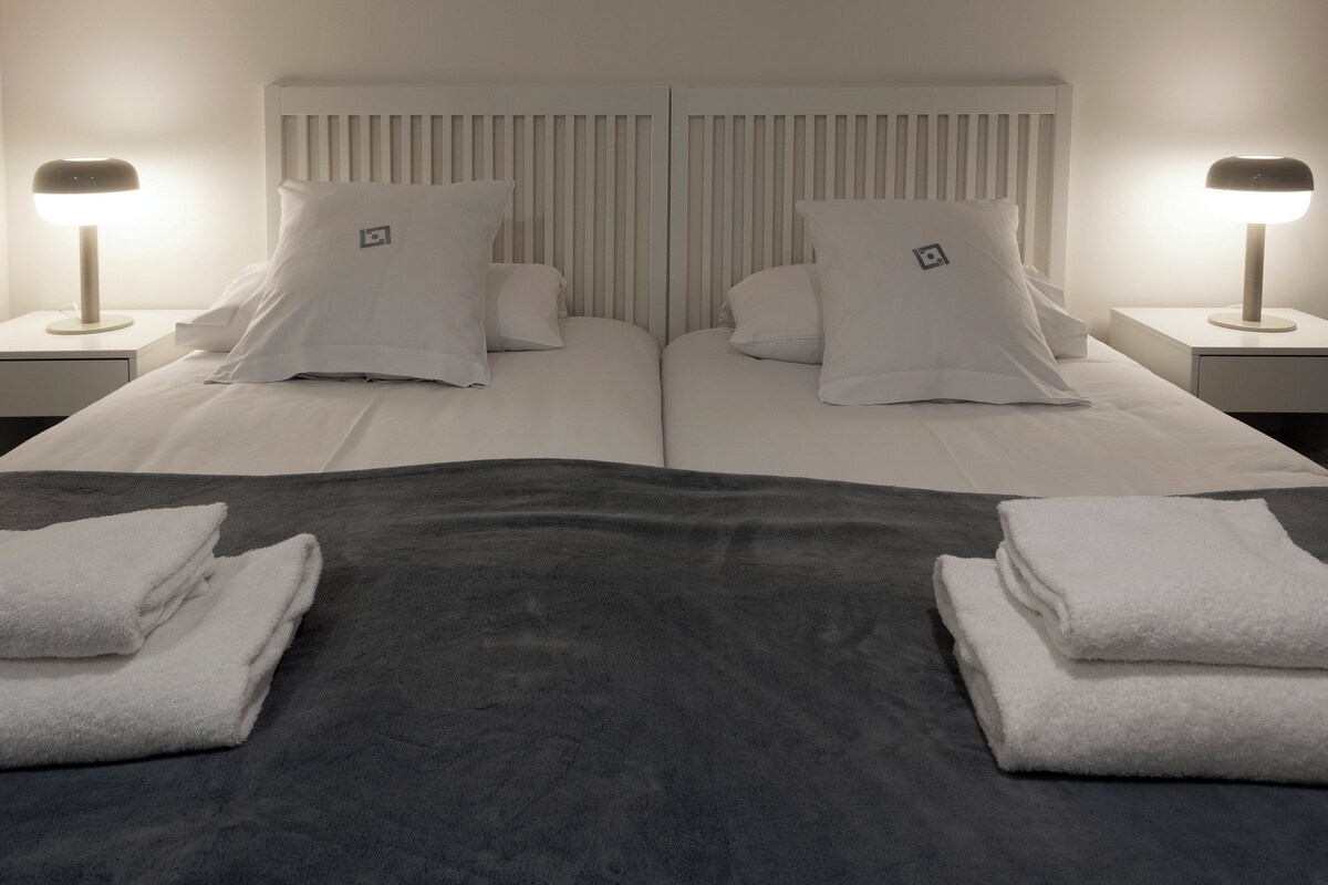 A double bed with white linens, two bedside tables with lamps, and folded towels on the bed.