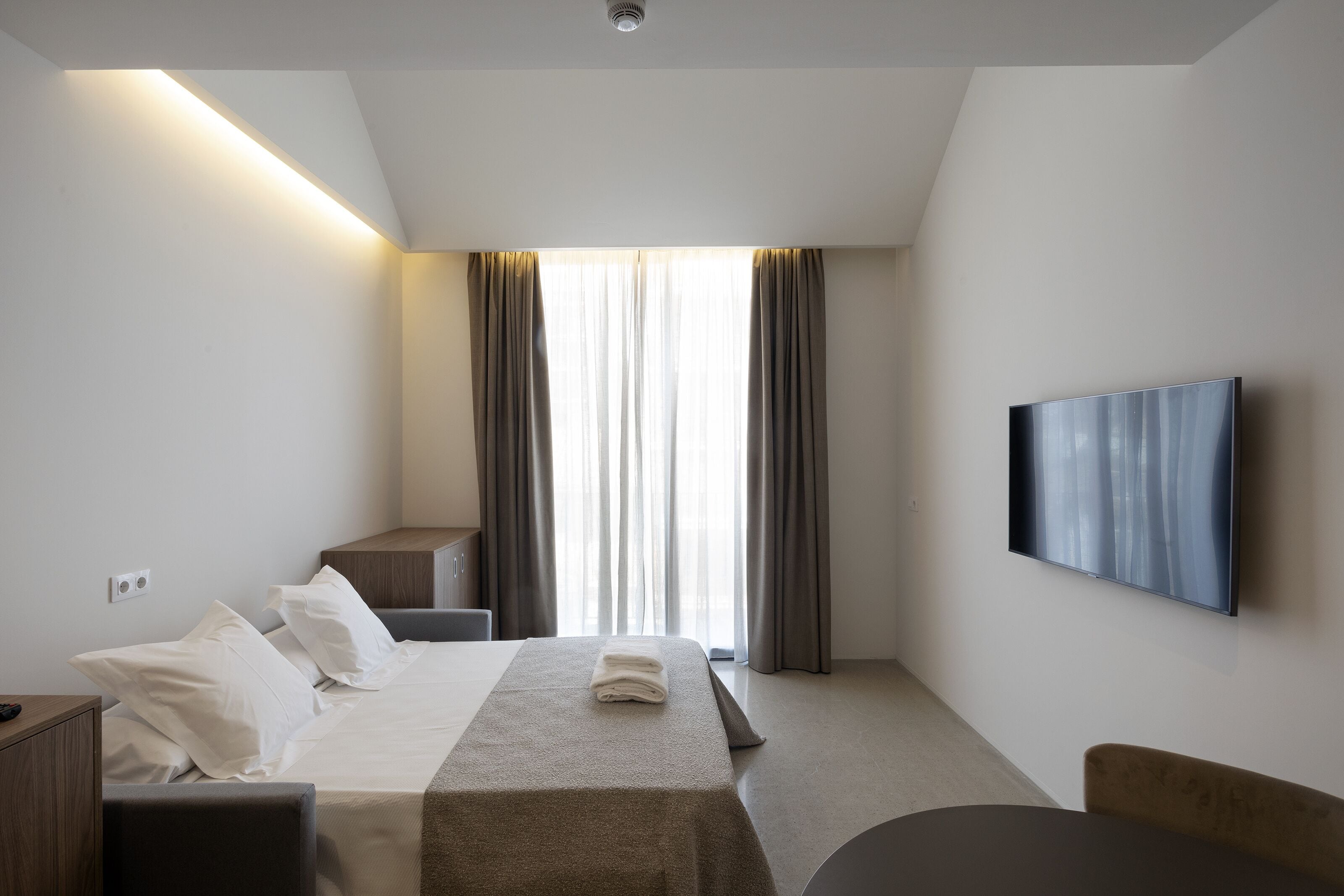 Habitación de hotel con cama, mesita de noche, silla, televisor y un ventanal con cortinas.