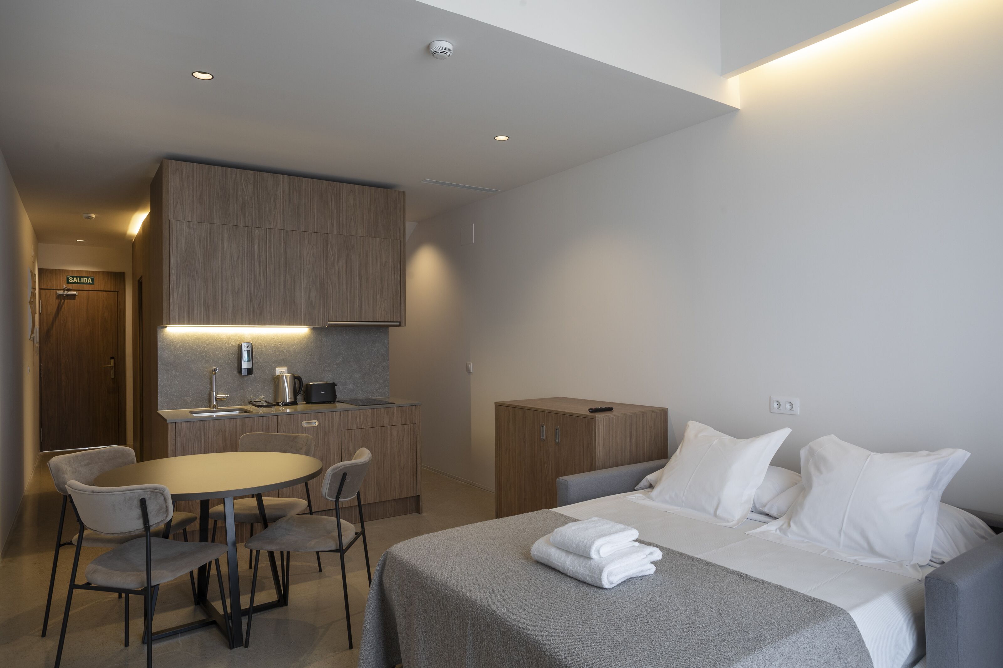 A modern hotel room with a bed, bedside table, and a small kitchenette.