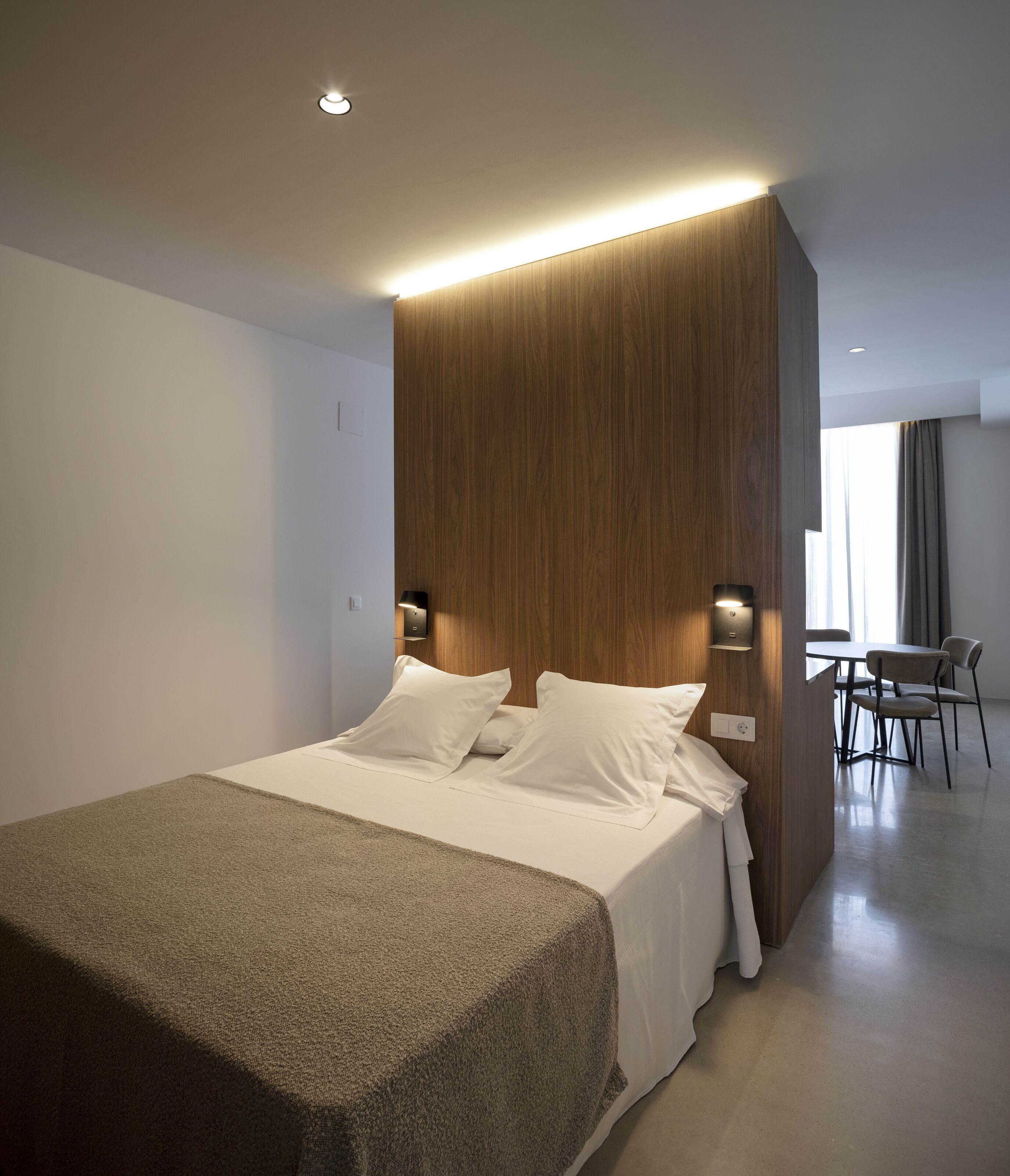 Une chambre d’hôtel moderne avec un grand lit, une tête de lit en bois, deux lampes de chevet et une vue sur l’espace repas.