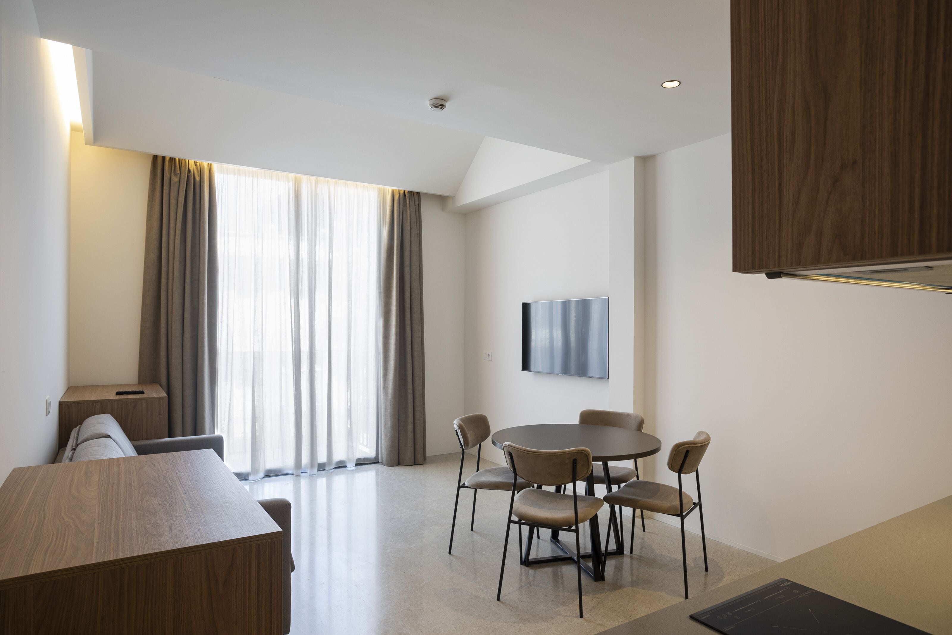 Een moderne hotelkamer met een houten bureau, een ronde eettafel met stoelen, een flatscreen televisie aan de muur en grote ramen met gordijnen.