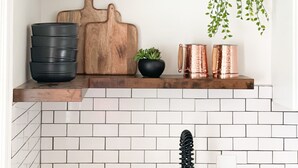 Una cocina con azulejos blancos tipo metro, una tabla de cortar de madera, un tazón negro y tazas de cobre en un estante.