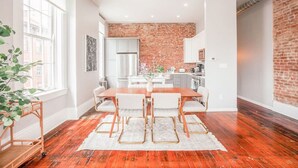 Une cuisine moderne avec des meubles blancs, un mur d’accent en briques et un coin repas comprenant une table et des chaises en bois.