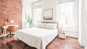 Une chambre avec un mur en briques, un lit, un bureau et de grandes fenêtres.