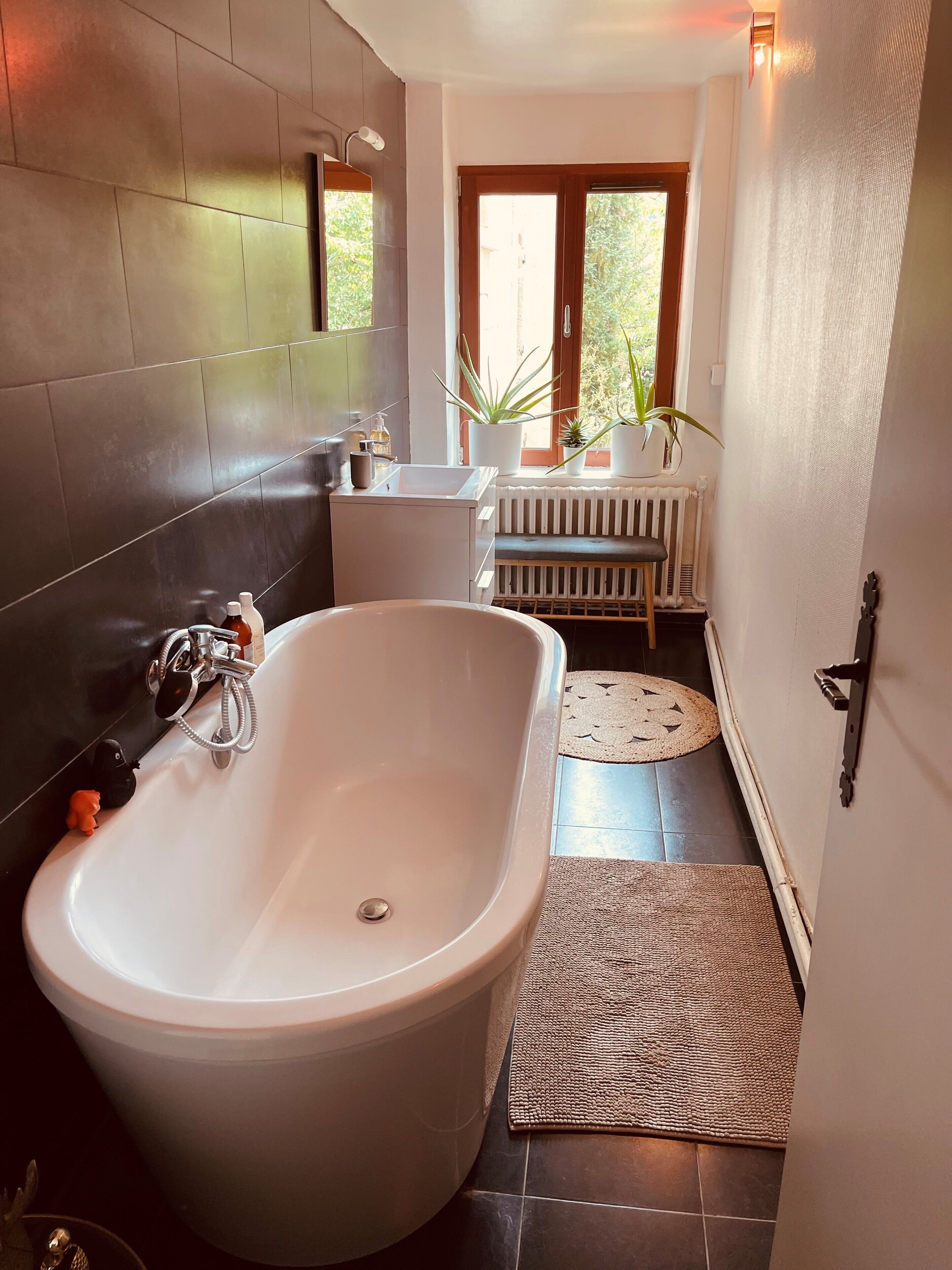 Ein modernes Badezimmer mit einer freistehenden Badewanne, einer Toilette, einem Waschbecken und einem Fenster mit Pflanzen.