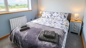 Une chambre à coucher avec un lit, deux tables de chevet avec des lampes, un radiateur et une fenêtre donnant sur un paysage verdoyant.