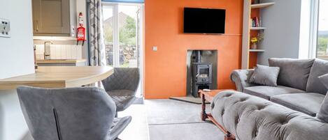A modern living room with a grey sofa, a wooden coffee table, and a grey armchair. A flat-screen TV is mounted on the orange wall, and a fireplace is visible in the background.
