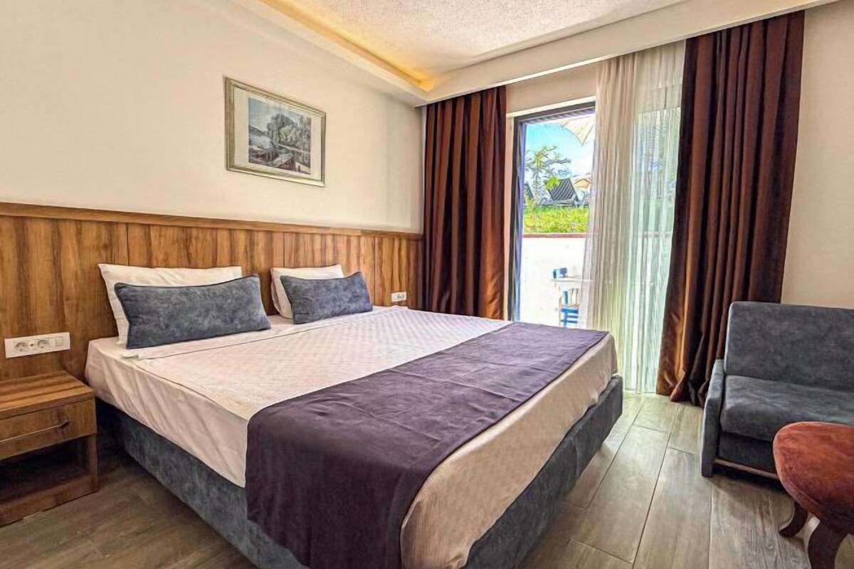 A bedroom with a bed, a nightstand, a chair, a window with curtains, and a balcony view.