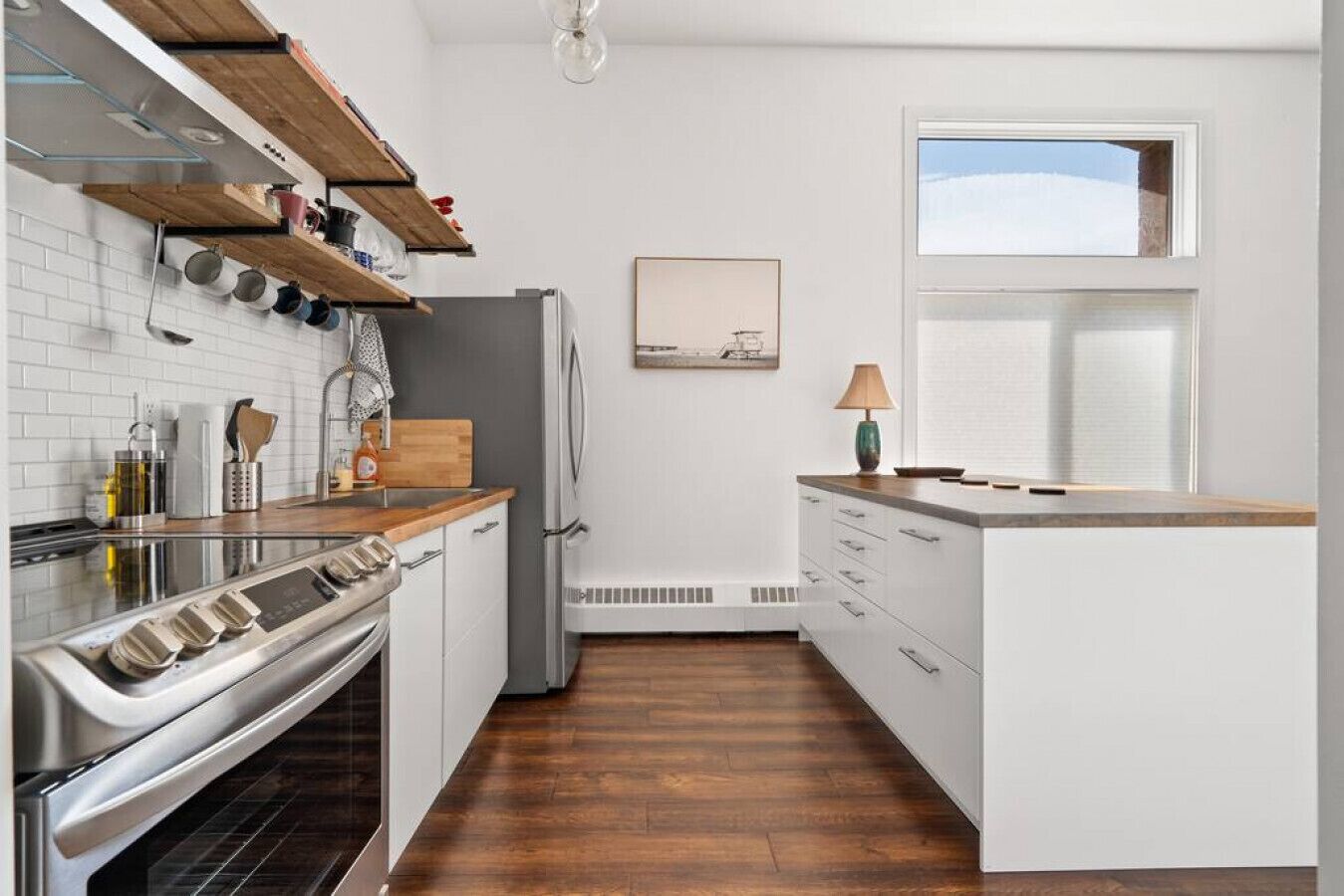 A modern kitchen with a built-in oven, wooden shelves, white cabinets, and a window with blinds.