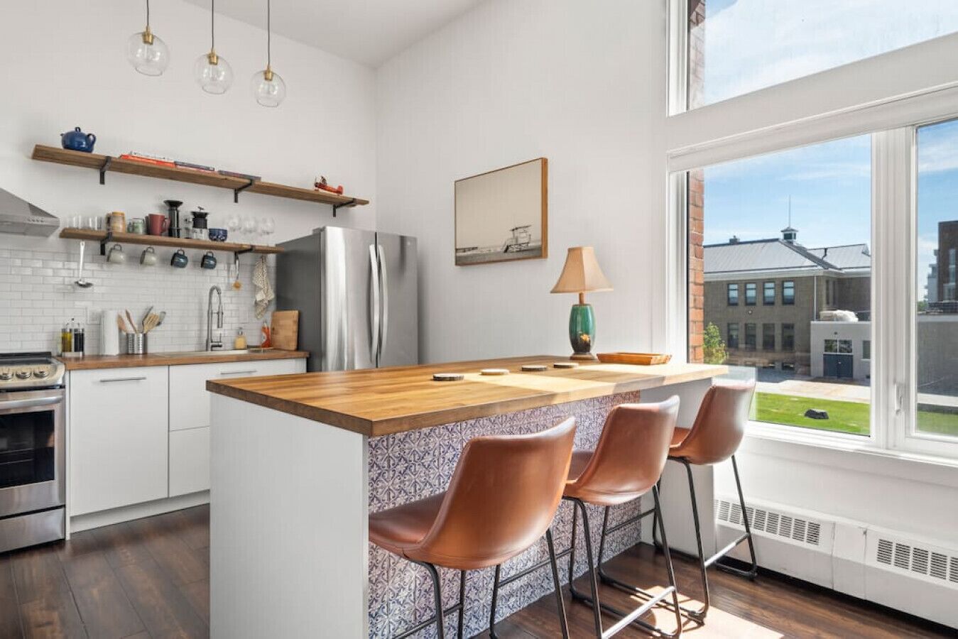 A modern kitchen with a central island, bar stools, and a view of a building through a large window.