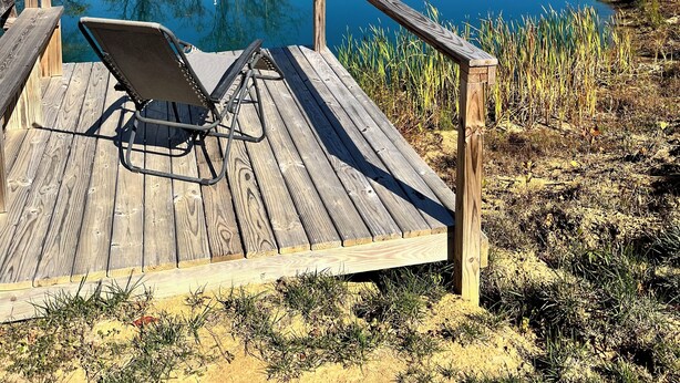 Terraza de madera con una silla y una escalera, con vista a un estanque azul y cristalino rodeado de árboles.