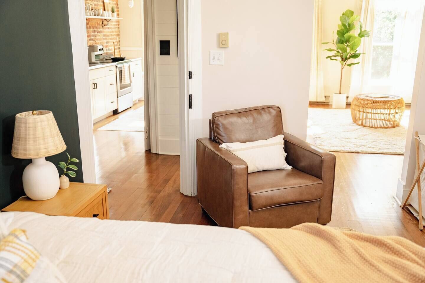Ett sovrum med en säng, en brun läderfåtölj, ett sängbord med en lampa och utsikt in i köket.