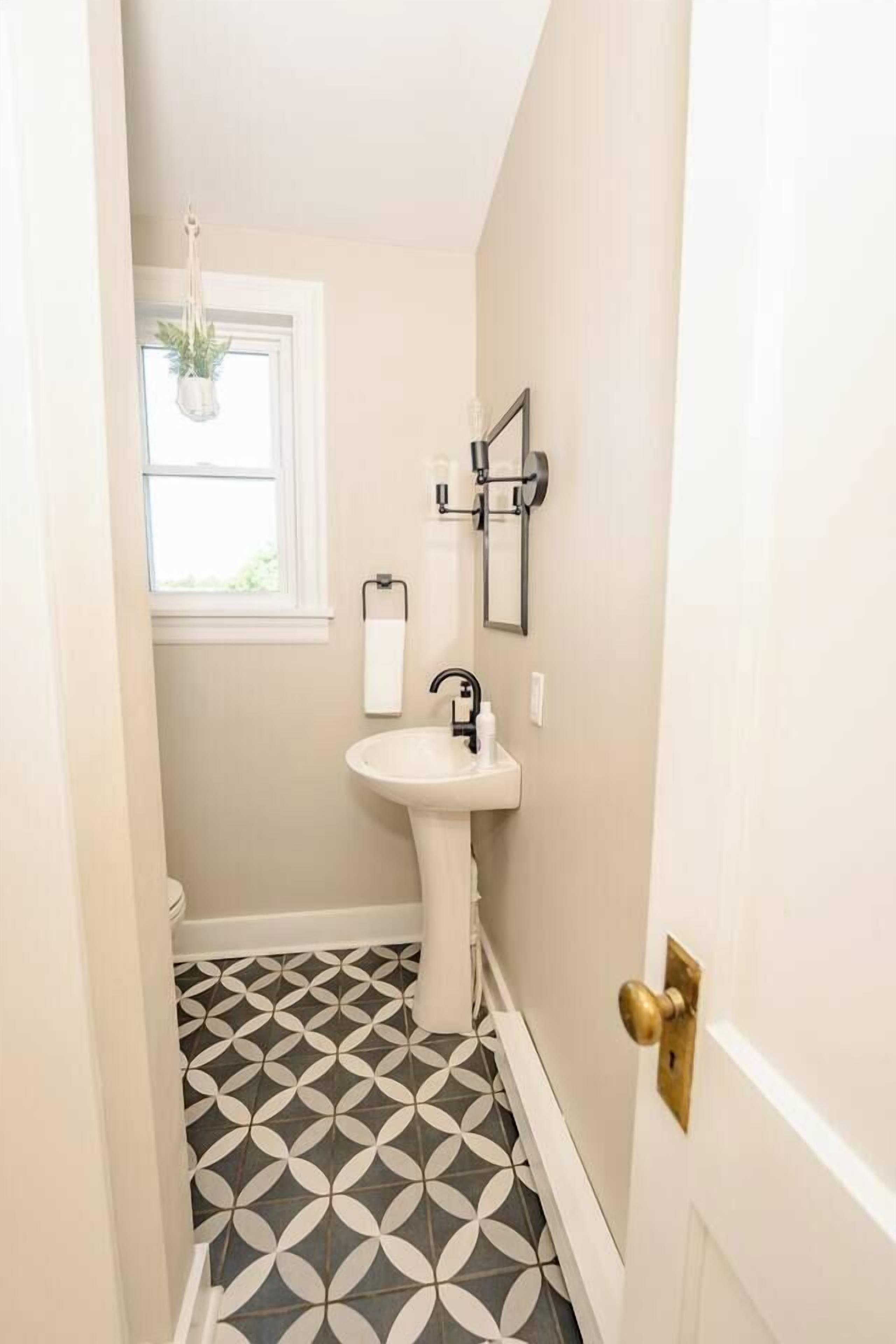 Un baño con un lavamanos blanco, una ventana con planta colgante y baldosas de suelo estampadas.