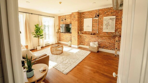 A spacious living room with a brick wall, a flat-screen TV, a large window, and a cozy seating area with a round table and chairs. - The Boho Penthouse on Main (Chambersburg)