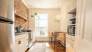 A kitchen with a brick wall, stainless steel appliances, a wooden dining table with chairs, and hanging pendant lights. - The Boho Penthouse on Main (Chambersburg)