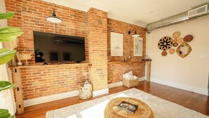 A living room with a brick fireplace, a flat-screen TV, and a decorative wall with various items. - The Boho Penthouse on Main (Chambersburg)