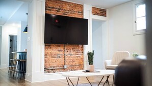 Uma sala de estar moderna com parede de tijolos decorativa, uma televisão de tela plana montada acima, uma mesa de centro redonda e uma área de estar confortável com uma poltrona e um sofá.