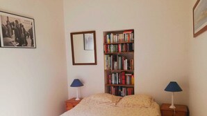 Ein Schlafzimmer mit einem Bett, einem Bücherregal, zwei Nachttischlampen, einem Spiegel und einem gerahmten Bild an der Wand.
