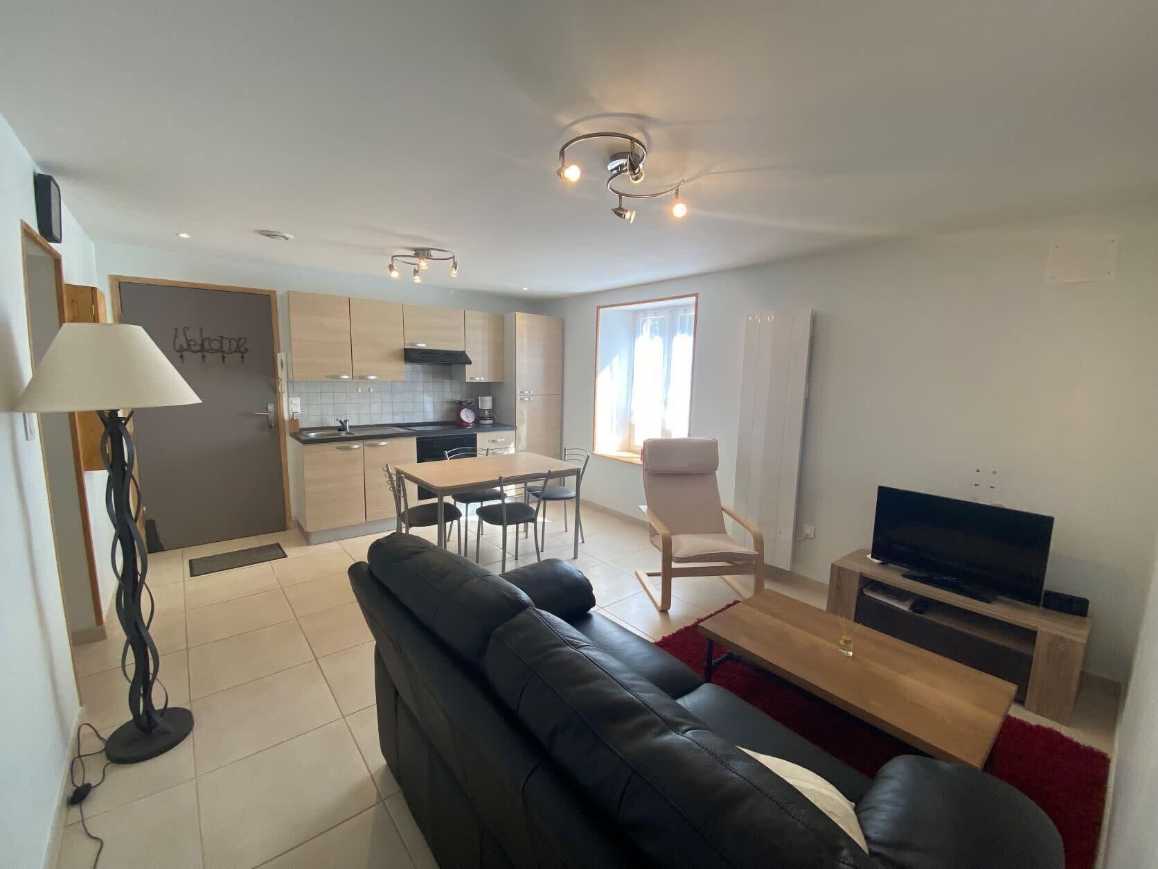A modern apartment with a living room, dining area, and kitchen. There is a black leather sofa, a dining table with chairs, a TV, and a lamp.