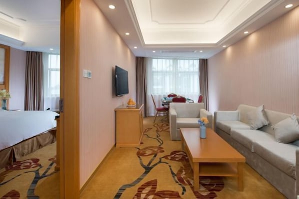 A hotel room with a bed, a sofa, a coffee table, a TV, and a dining area with a table and chairs.