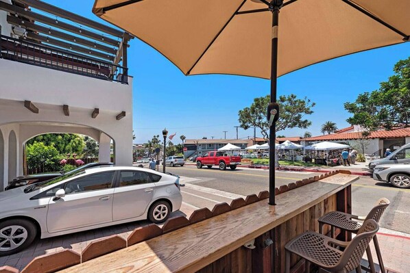 A white car parked in front of a building with a balcony, a wooden bench, and outdoor seating with umbrellas.