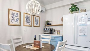 A modern kitchen with a white refrigerator, a coffee machine, and a dining table with chairs. There are framed botanical prints on the wall and a hanging light fixture. - Quiet Condo in Village with Deck, Bikes and AC (San Clemente)