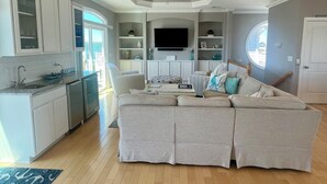 A spacious living area with a large sectional sofa, a kitchen with white cabinetry, and a ceiling fan. - Beach House w/beautiful ocean views.  Private heated pool. Elevator. Sleeps 10. (Ocean Isle Beach)