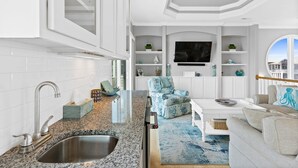 A modern kitchen with a granite countertop, stainless steel sink, and white cabinetry. There is a blue floral armchair and a white sofa with a blue rug in the living area. - Beach House w/beautiful ocean views.  Private heated pool. Elevator. Sleeps 10. (Ocean Isle Beach)