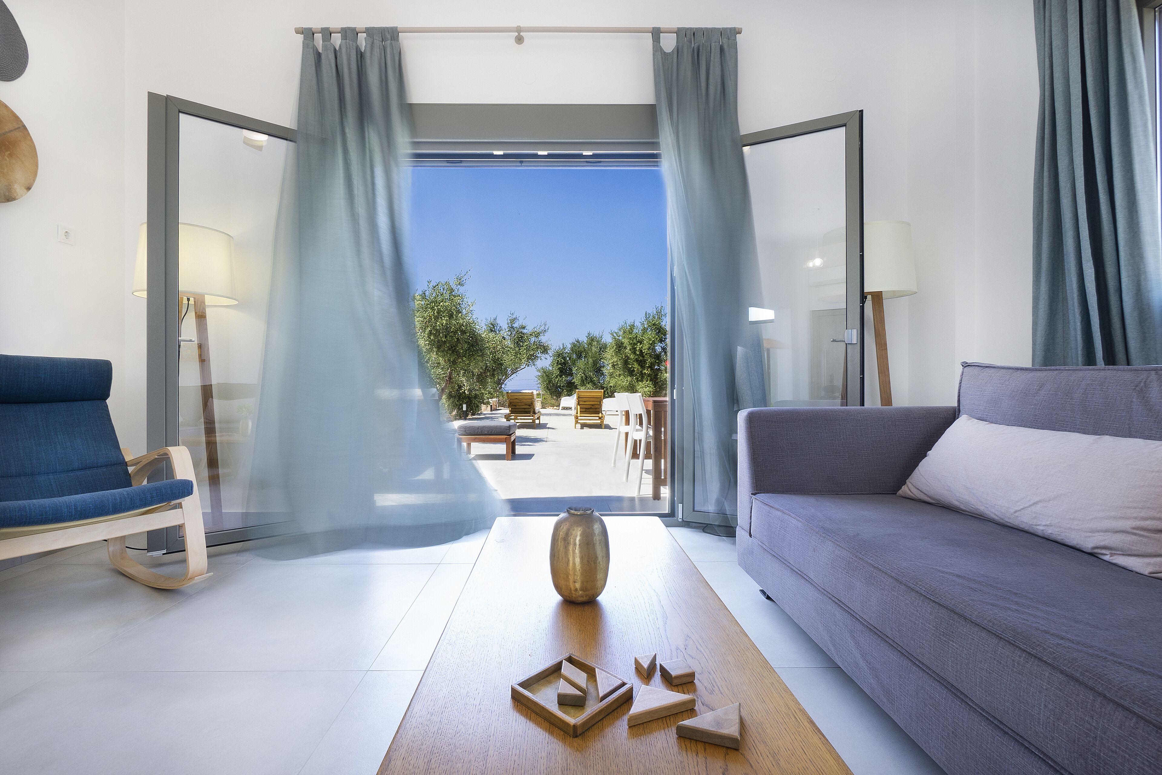 A modern living room with a sofa, a chair, a coffee table with geometric wooden sculptures, and a view of an outdoor patio with furniture.