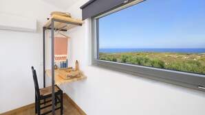 Una habitación con un ventanal grande con vistas a un paisaje costero, una silla de madera, una mesita con un juego de mesa y un estante colgante con una cesta y una toalla.