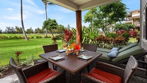 Une terrasse extérieure aménagée avec des meubles en osier et une table dressée pour les repas, le tout entouré d’une végétation luxuriante et de palmiers.