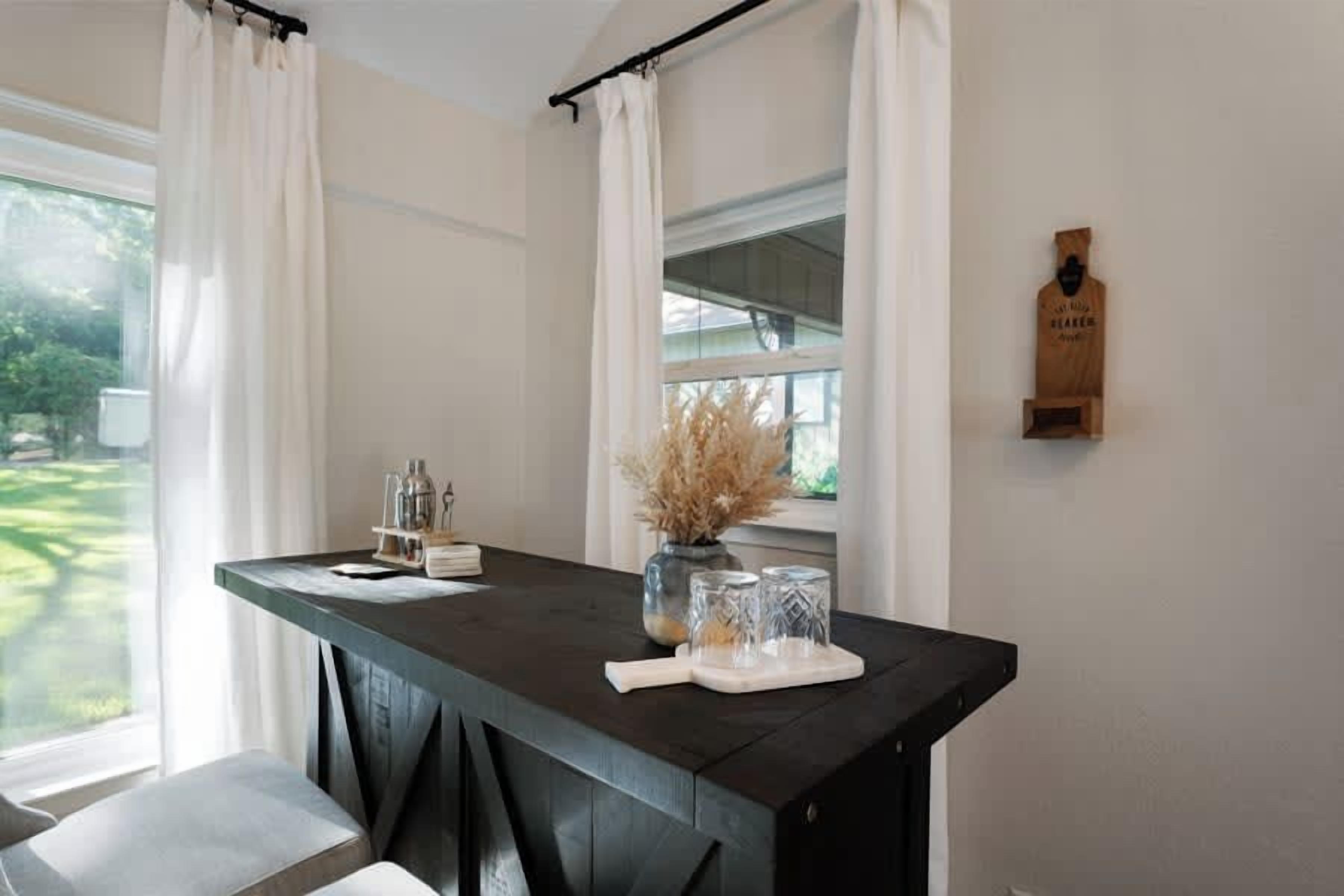 A room with a dark wooden table, white curtains, a window with a view of greenery, and a wooden wall-mounted object.