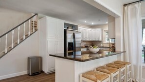 A modern kitchen with white cabinets, a stainless steel refrigerator, a central island with bar stools, and a staircase leading to another level.