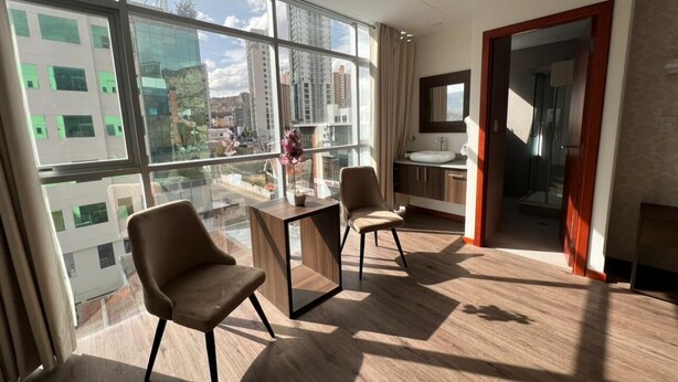 A modern hotel room with large windows, a wooden floor, two chairs, a small table, and a bathroom visible through an open door. - NOVUM HOTEL (La Paz)