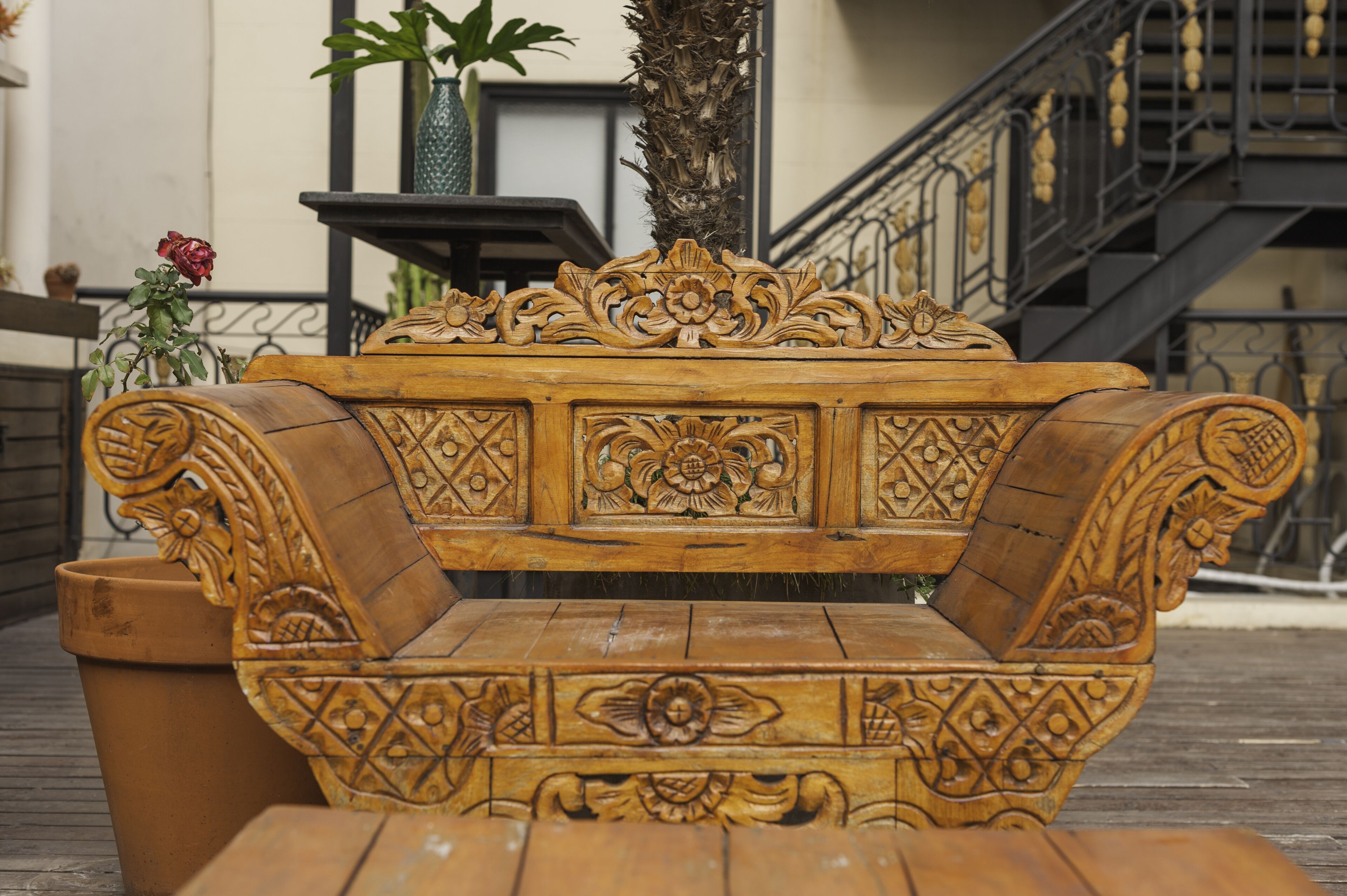 A wooden bench with intricate carvings, a potted plant, and a staircase in the background.